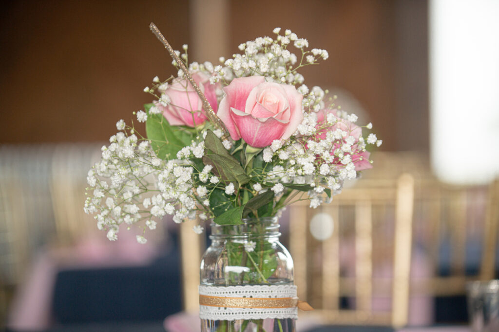 Mason jar centerpiece with pink roses, baby’s breath, greenery, and lace ribbon, styled on a table set for an event.