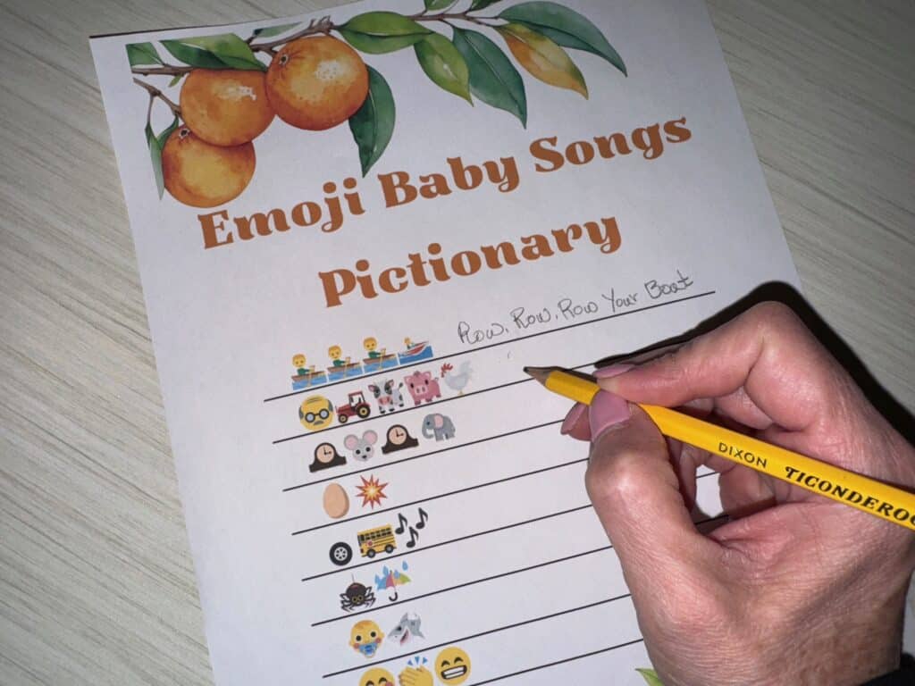 Hand holding a pencil while filling out an Emoji Baby Songs Pictionary game sheet, featuring rows of emoji clues and space to write in the song titles.