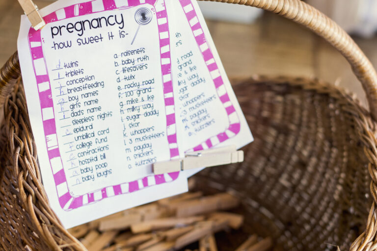 Baby shower game setup featuring a wicker basket filled with wooden clothespins and printed “How Sweet It Is” candy‑matching game sheets with a purple striped border.