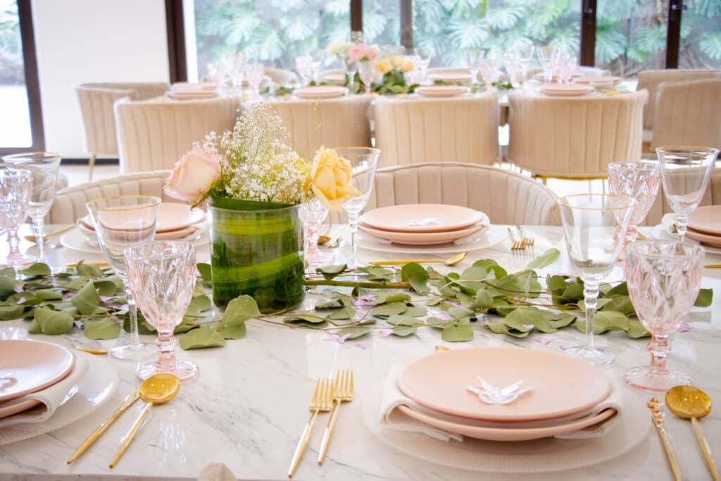 A beautifully styled baby shower tablescape featuring a white table with a lush greenery garland, a floral arrangement in a green vase, pink plates with butterfly accents, gold flatware, and pink goblets, set in a bright room with large windows and soft neutral chairs.