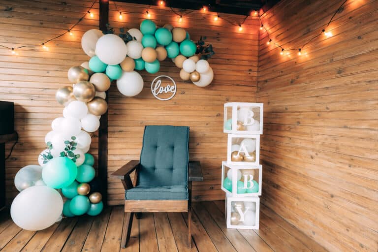 A cozy indoor baby‑shower setup featuring a teal armchair centered beneath a balloon arch in white, gold, and teal tones, with a circular “love” sign behind it and stacked BABY boxes to the side, creating a clear focal point for photos.