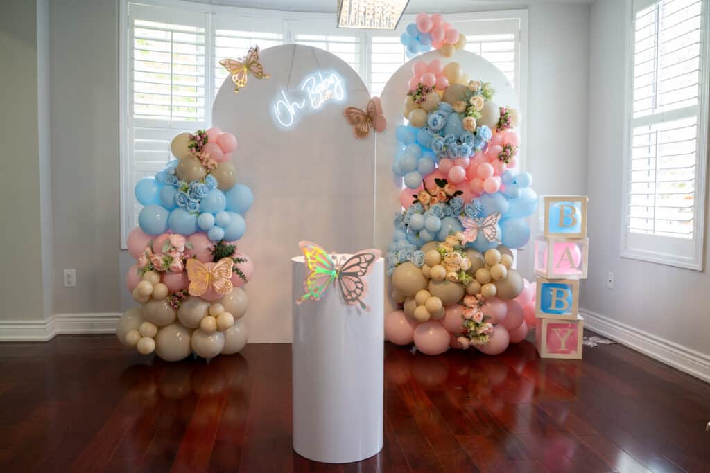 An indoor baby‑shower backdrop with pastel balloon arrangements framing a white arch that reads “Oh Baby,” styled with butterflies, a pedestal, and BABY letter boxes, all arranged with open floor space for guests to move comfortably around the setup.