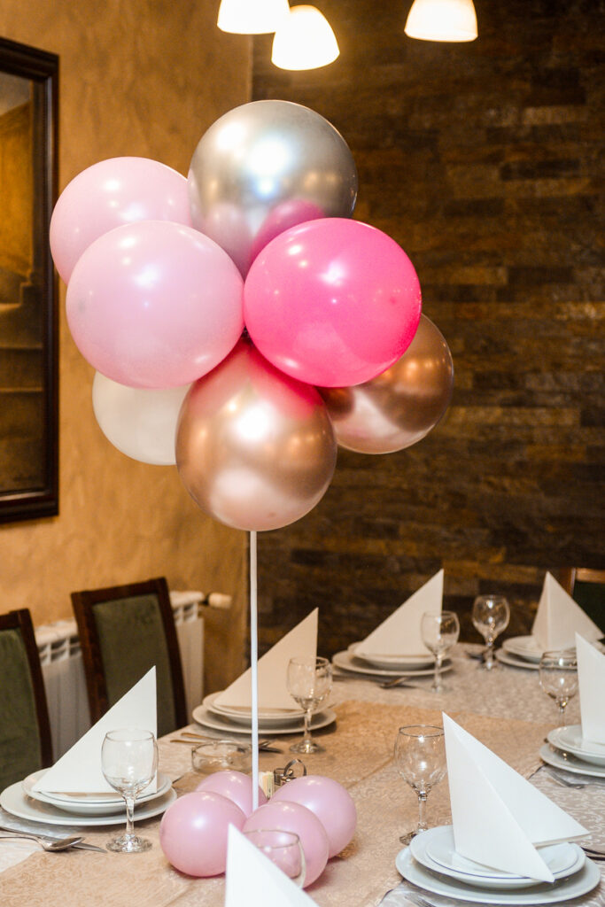 Balloon centerpiece with pink, white, and silver balloons arranged on a dining table set with plates, folded napkins, and glassware for a baby shower.