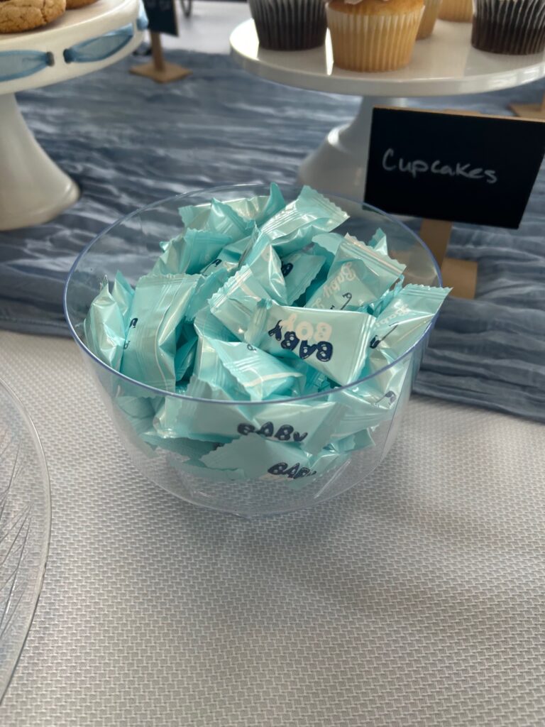 A clear bowl filled with individually wrapped baby‑themed mints displayed on a table beside a stand of cupcakes.