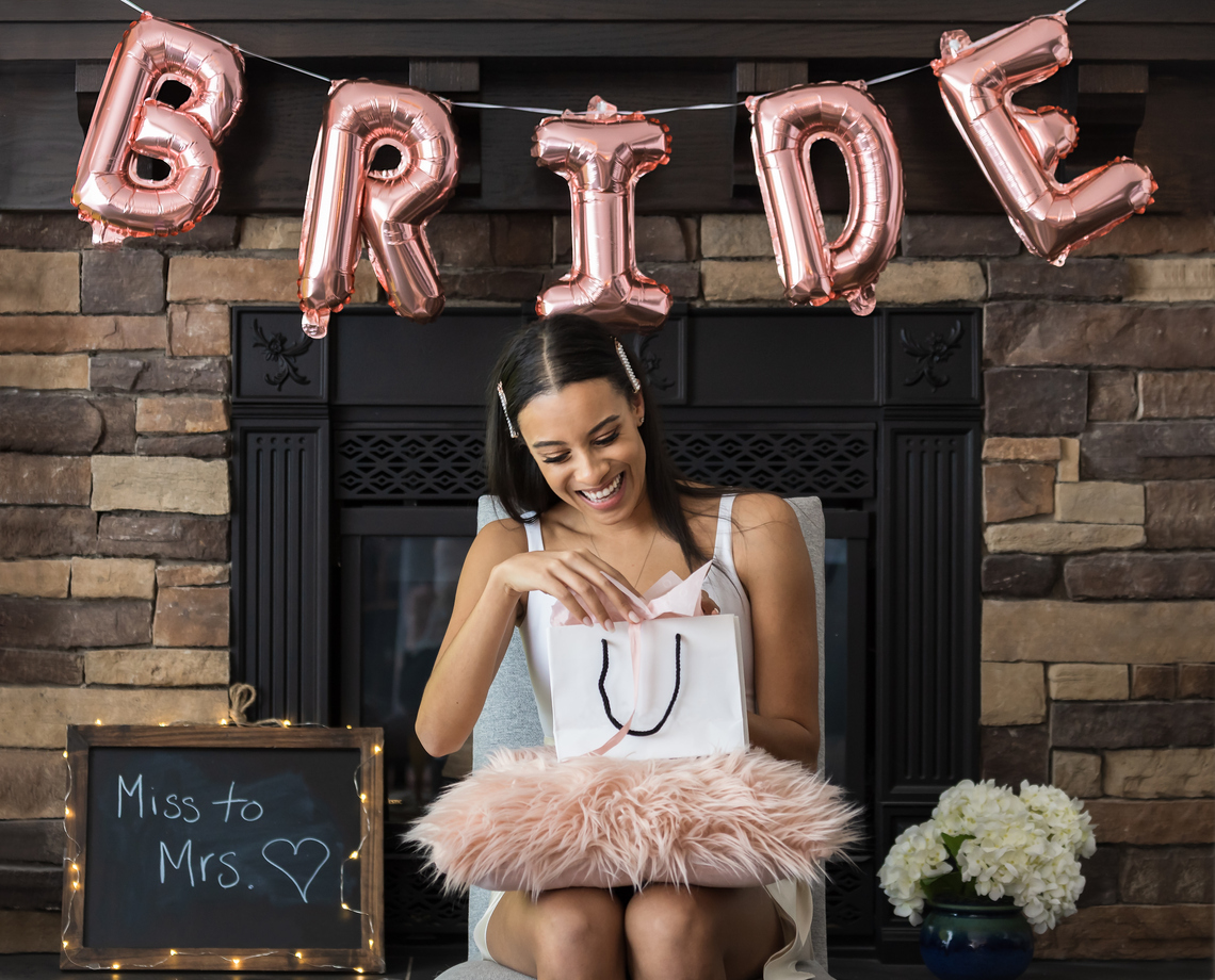 woman opening gift with a garland of ballons spelling 'bride" behind her