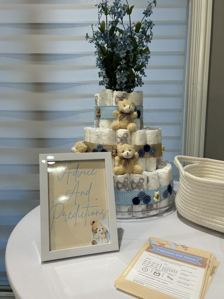 Baby shower advice and predictions table featuring a multi‑tiered diaper cake, a repurposed white frame displaying a themed sign, and a woven basket used to hold cards and supplies.