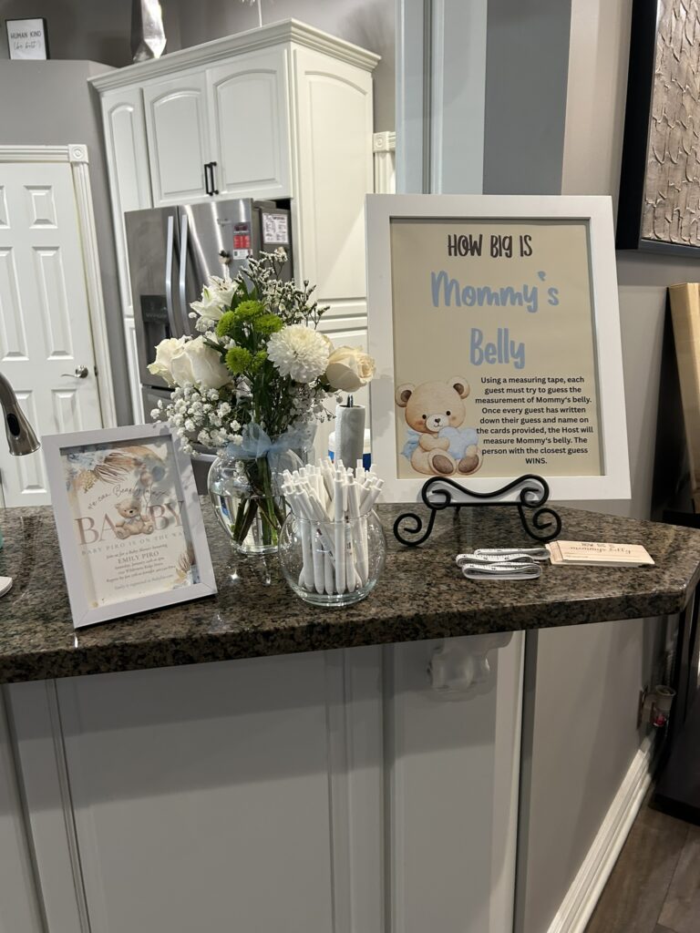 Baby shower advice and predictions table featuring a tiered diaper cake decorated with teddy bears and blue accents, alongside a repurposed white frame displaying a themed sign and a woven basket holding cards.