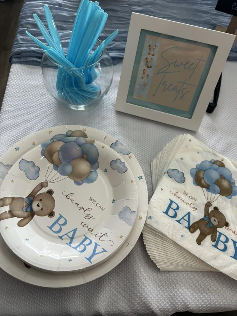 Glass bowl of blue plastic utensils next to a framed “Sweet Treats” sign and matching bear‑themed plates and napkins in blue and white.