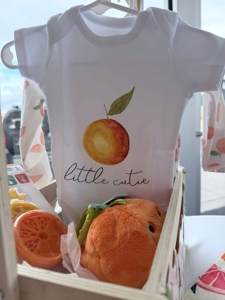 A wooden gift crate displaying a baby onesie with an orange design, a plush orange toy, a teething toy, and other citrus‑themed baby items.