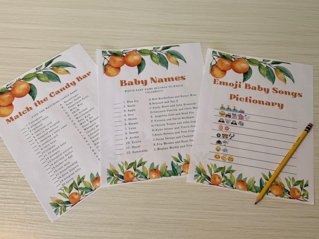 A flat lay of three Little Cutie–themed baby shower game cards on a light wood surface, including Match the Candy Bar, Baby Names in Songs, and Emoji Baby Songs Pictionary, with a pencil placed beside them.