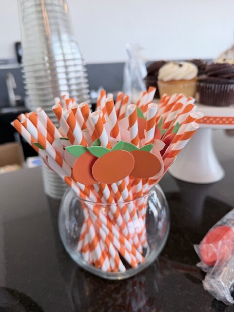 Orange‑themed paper straws in a glass container, decorated with small citrus cutouts and displayed beside cupcakes and cups.
