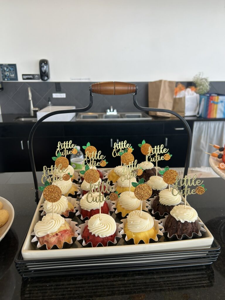 Tray of assorted mini bundt cakes topped with “Little Cutie” orange‑themed picks, arranged in a white tray with a black handle on a kitchen counter.