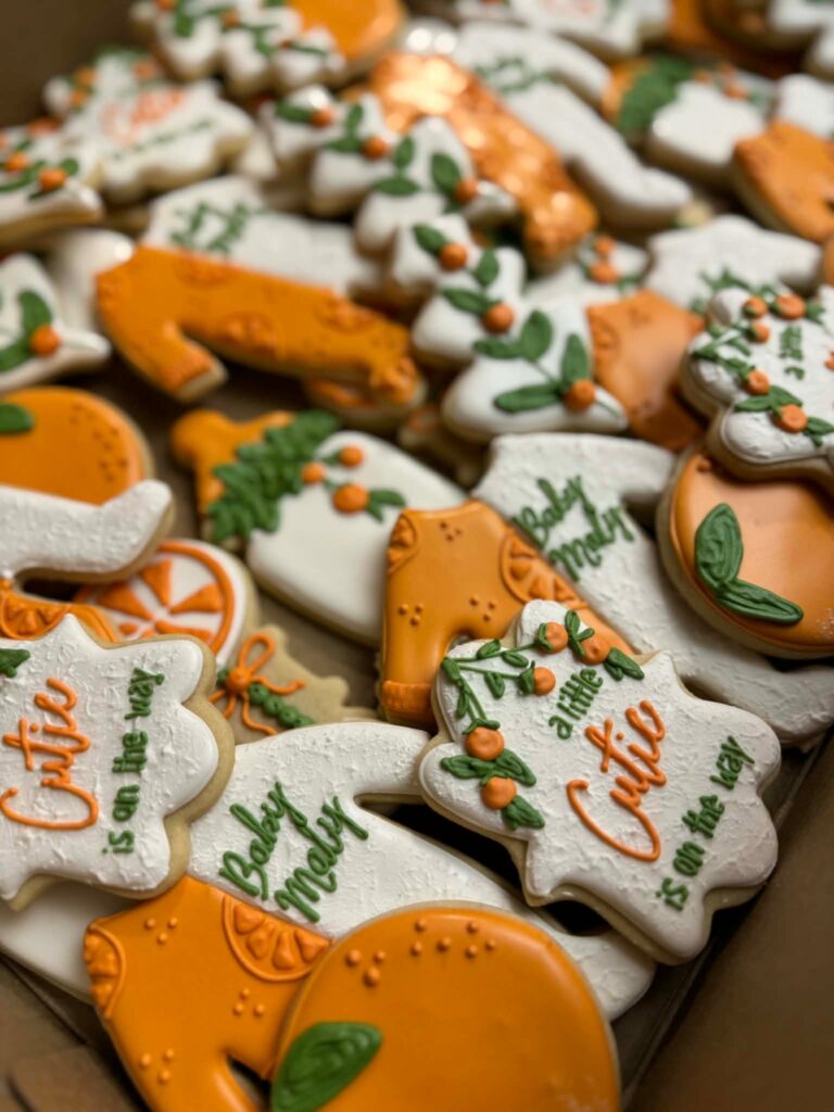 Decorated Little Cutie baby shower cookies arranged together, featuring oranges, greenery, baby onesies, bottles, and themed messages in orange, green, and white icing.