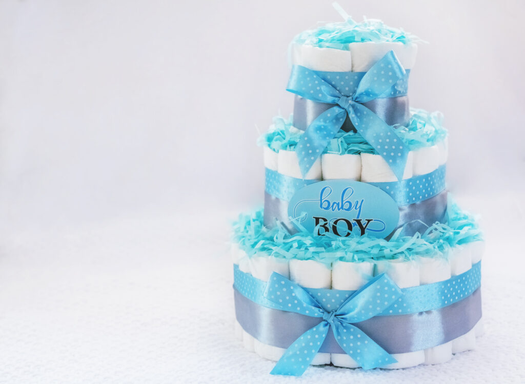 Three‑tier diaper cake decorated with blue and silver ribbon, shredded paper, and a “baby boy” sign, styled as a baby shower centerpiece.