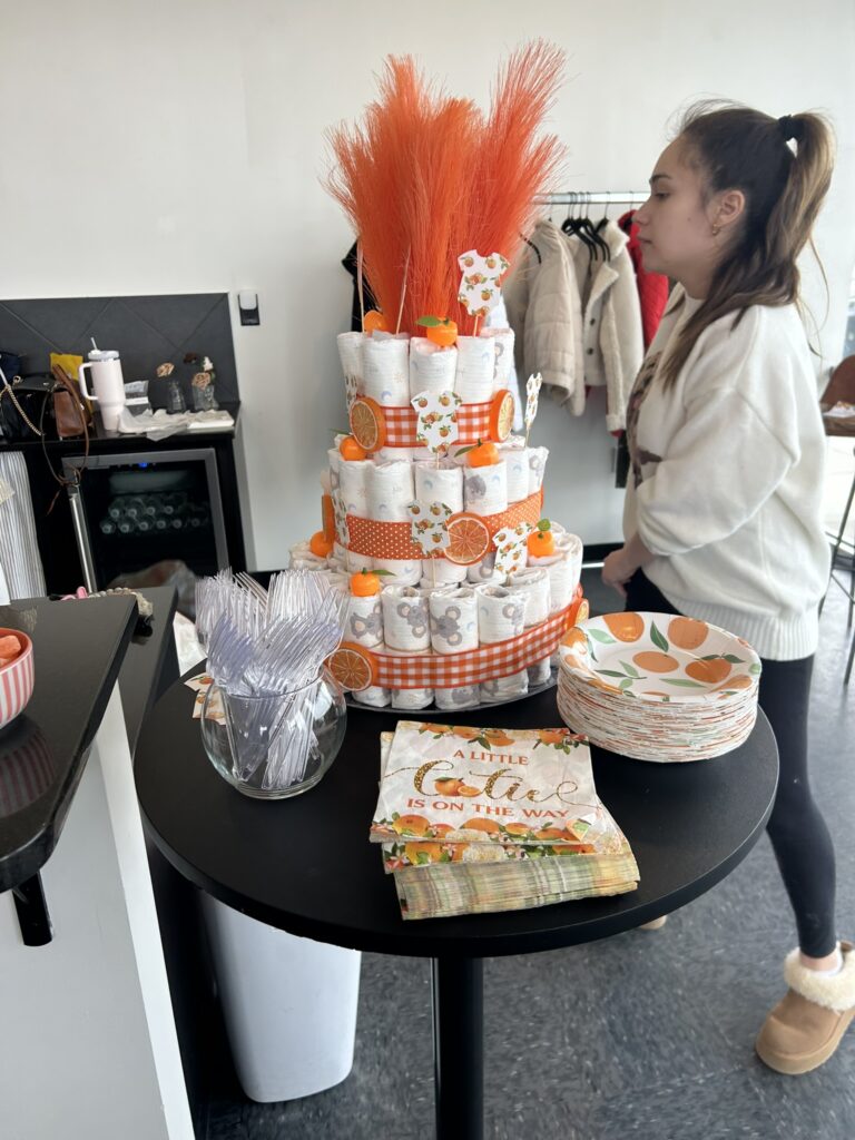 A three‑tier diaper cake decorated with orange slices, small faux oranges, and orange ribbon, displayed on a round table with matching plates, napkins, and utensils for a Little Cutie baby shower.
