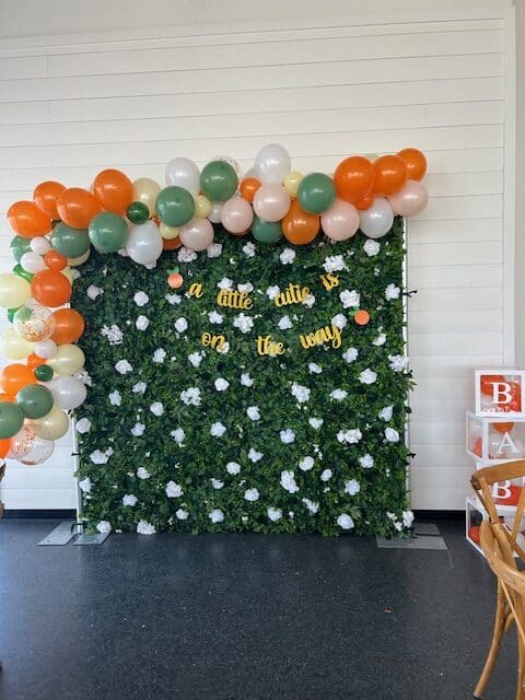 A baby shower backdrop featuring a lush green faux‑foliage wall with white flowers and yellow letters spelling “a little cutie is on the way.” A colorful balloon garland in shades of orange, green, white, peach, and clear confetti balloons frames the setup. Clear display boxes stacked on the right spell “BABY,” with orange items inside, and a wooden chair sits nearby.