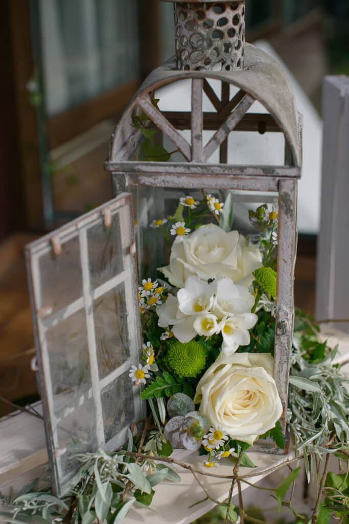 A rustic, weathered lantern filled with white roses, freesias, daisies, and soft greenery, styled as a baby shower centerpiece with additional foliage surrounding the base.