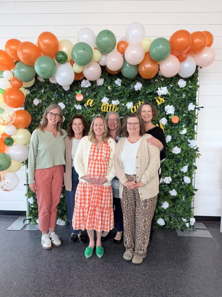A group of guests standing in front of a Little Cutie baby shower backdrop with a greenery wall, orange and sage balloon garland, and a banner that reads “a little cutie is on the way.