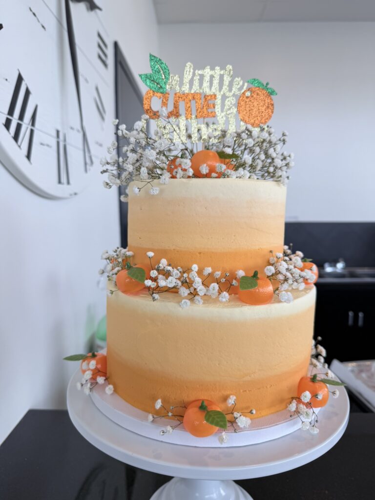 A two‑tier Little Cutie baby shower cake decorated with orange and cream frosting, small faux oranges, greenery, and a glittery “a little cutie is on the way” topper.