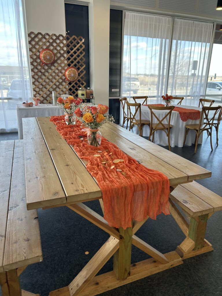 A wooden picnic-style table decorated for a Little Cutie baby shower with an orange table runner, dried citrus slices, and small floral centerpieces, with additional citrus-themed tables and décor in the background.