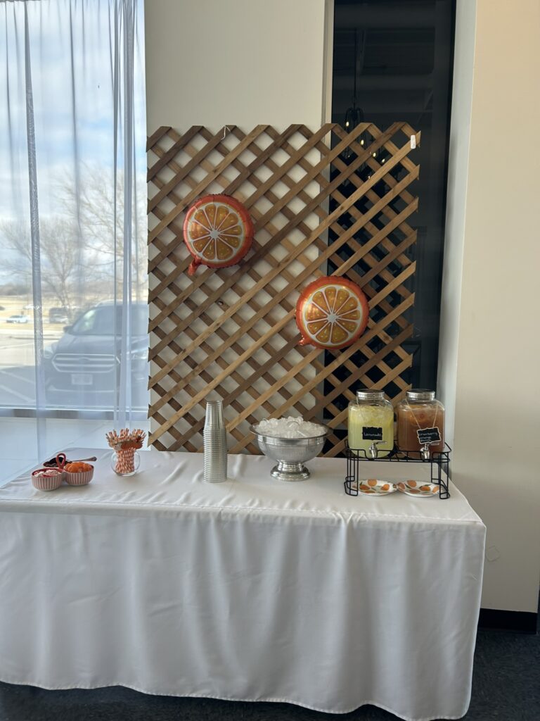 A citrus‑themed drink station with lemonade and sweet tea dispensers, orange slice plates, striped straws, and small bowls of garnishes displayed on a white table.