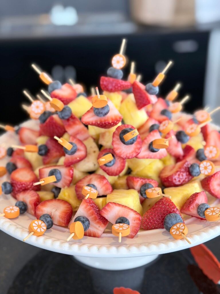 Fruit kabobs arranged on a white pedestal plate, made with fresh berries, pineapple, and banana, each topped with a small orange-themed skewer for a Little Cutie baby shower.