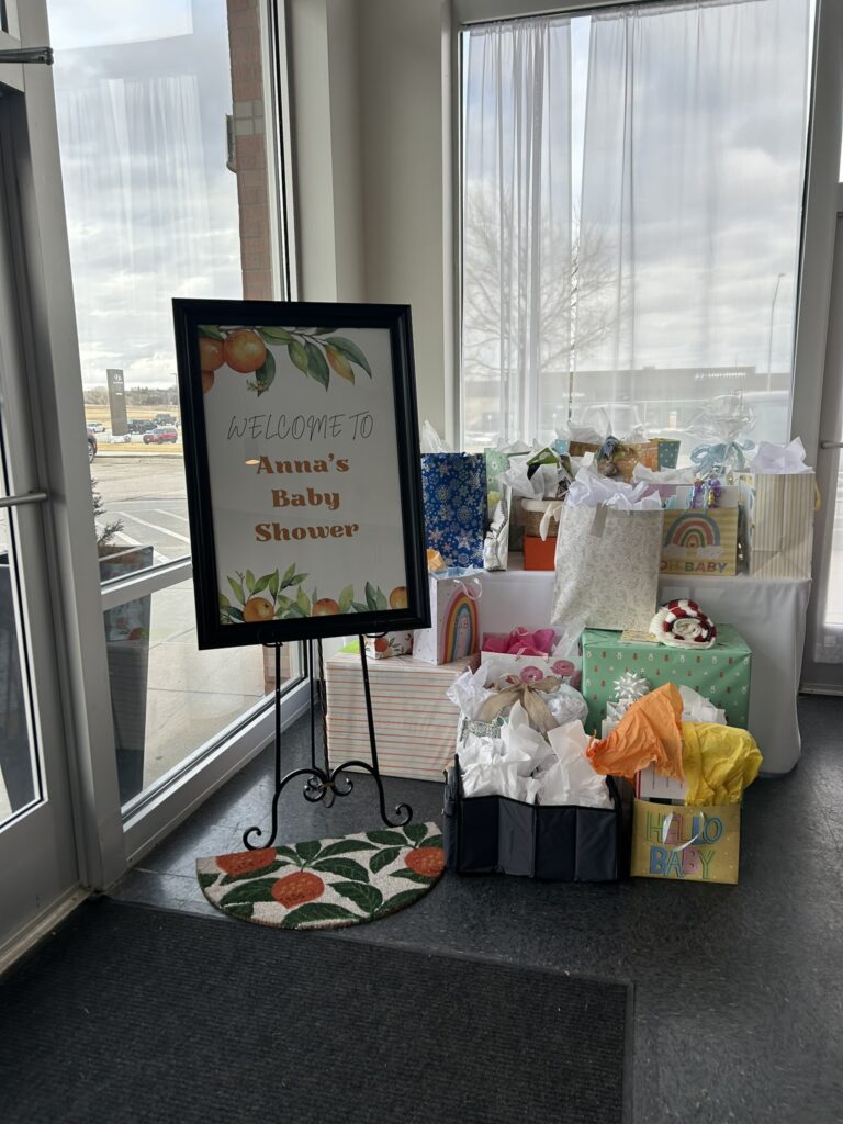 A baby shower gift table with a framed welcome sign featuring citrus accents, surrounded by pastel gift bags and boxes arranged near a bright window.