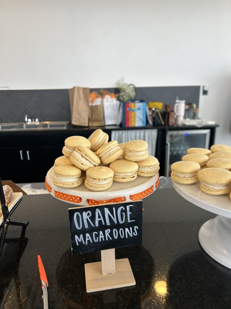 Orange macarons displayed on two cake stands, with a small chalkboard sign labeled “orange macarons” and a polka‑dot ribbon accent for a Little Cutie baby shower.