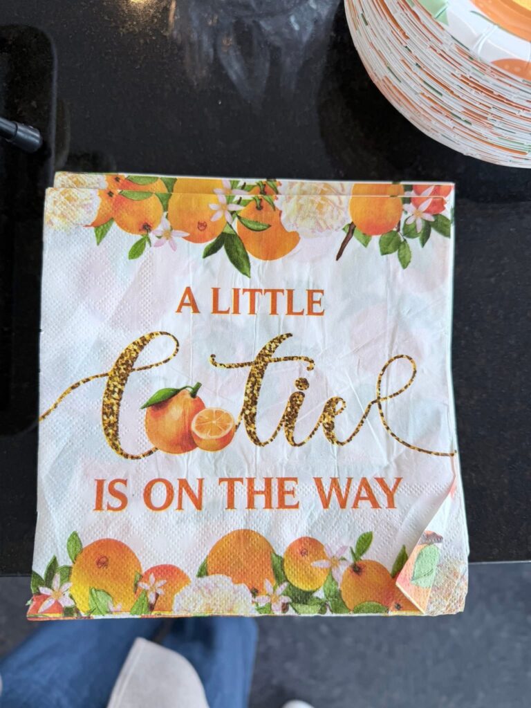 A Little Cutie baby shower napkin decorated with oranges, blossoms, and greenery, featuring a glittery “cutie” design with matching themed plates nearby.