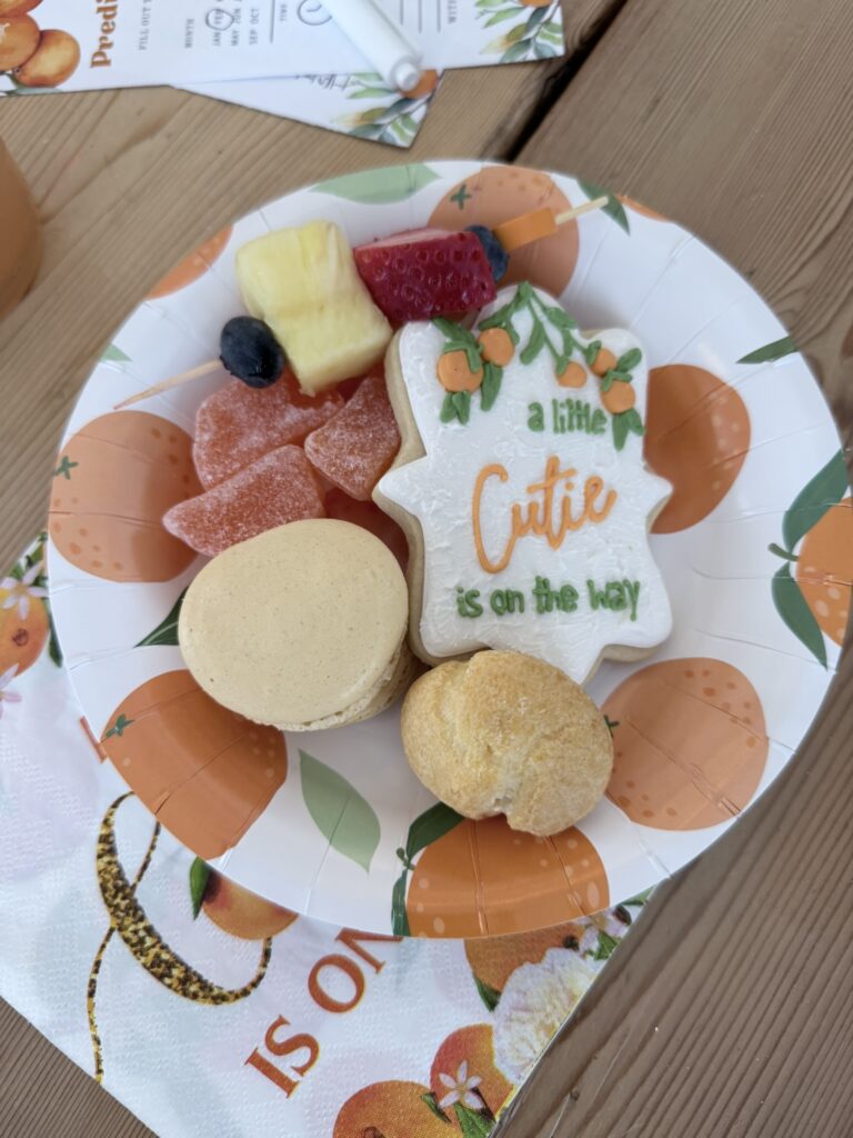 A themed paper plate filled with baby shower treats, including a decorated cookie, fruit skewer, macaron, gummies, and a matching orange‑print napkin underneath.