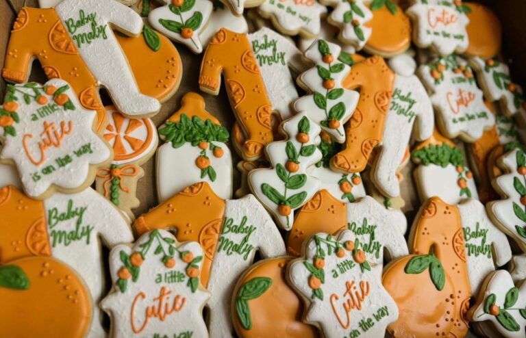 Decorated sugar cookies for a Little Cutie baby shower, including onesies, bottles, oranges, and themed plaques in orange, green, and white icing.
