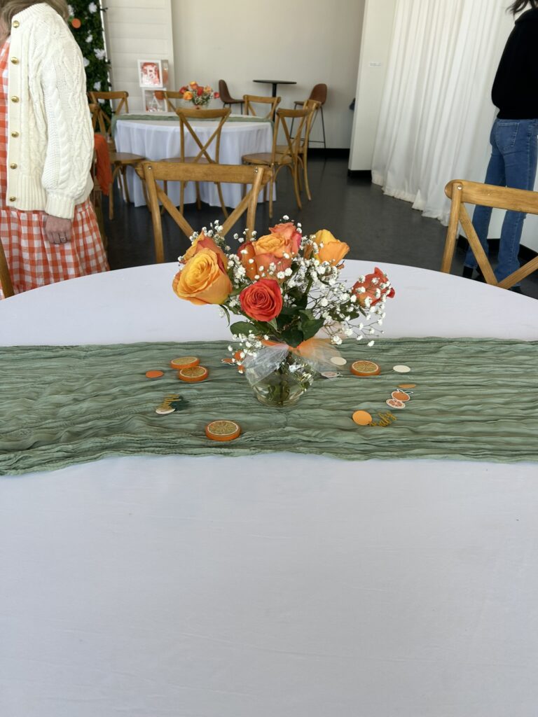 A round table decorated for a Little Cutie baby shower with a sage green table runner, dried orange slices, and a floral centerpiece featuring orange and red roses with baby's breath.