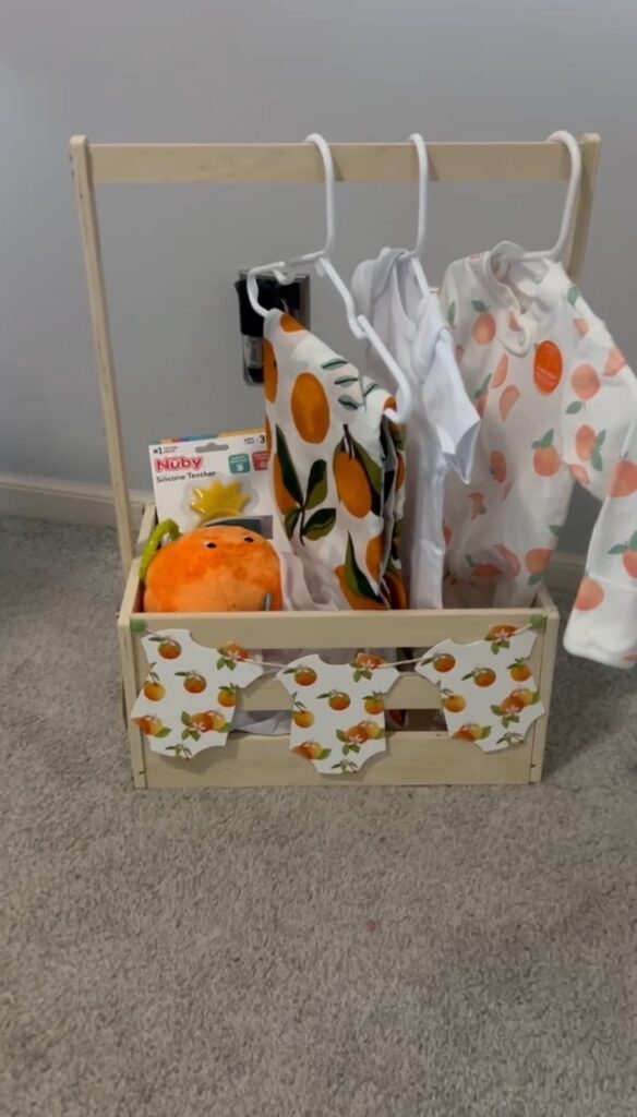 A wooden mini clothing‑rack gift basket holding baby outfits on hangers, with orange‑themed onesies, a plush orange toy, and baby items arranged inside the basket.