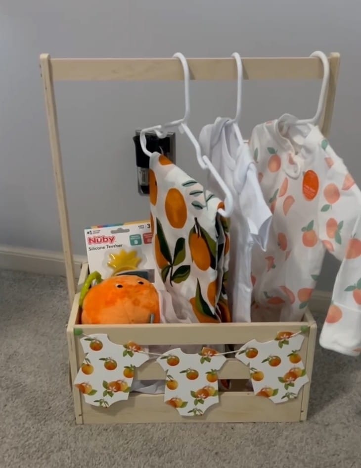 A wooden baby shower gift crate styled as a centerpiece, filled with fruit‑themed baby items including small outfits on hangers, a plush orange toy, a teether, and a mini onesie garland across the front, with soft natural light in the background.