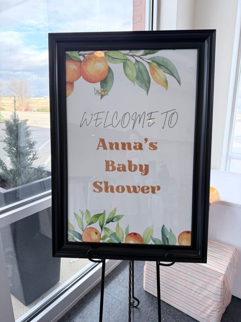 A framed Little Cutie baby shower welcome sign on an easel, decorated with illustrated oranges and greenery, displayed near a window at the party entrance.