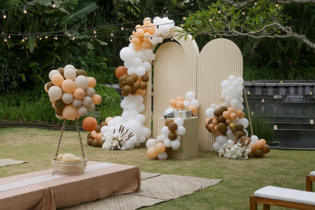 A layered outdoor baby‑shower backdrop featuring neutral arch panels, clustered balloons in warm earth tones, wrapped gifts on a pedestal, and soft string lights above, all styled on a grassy lawn surrounded by greenery.