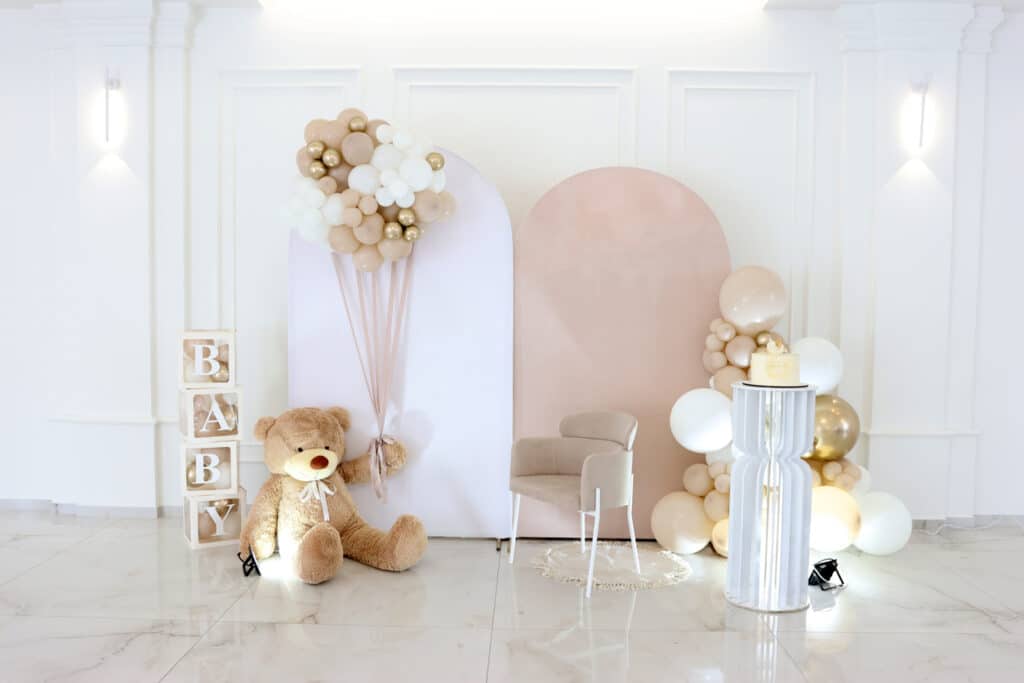 Baby shower photo spot featuring a blush and white backdrop, neutral balloon garland, a plush teddy bear beside stacked BABY blocks, a cozy chair on a round rug, and a small cake displayed on a white pedestal.