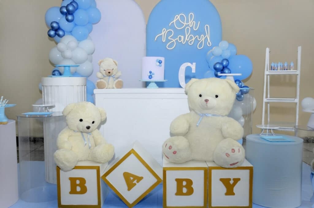 Baby‑shower backdrop in blue and white with balloon clusters, an “Oh Baby!” sign, a small cake on a white table, teddy bears sitting on BABY blocks, and coordinated props that all match the soft blue theme.