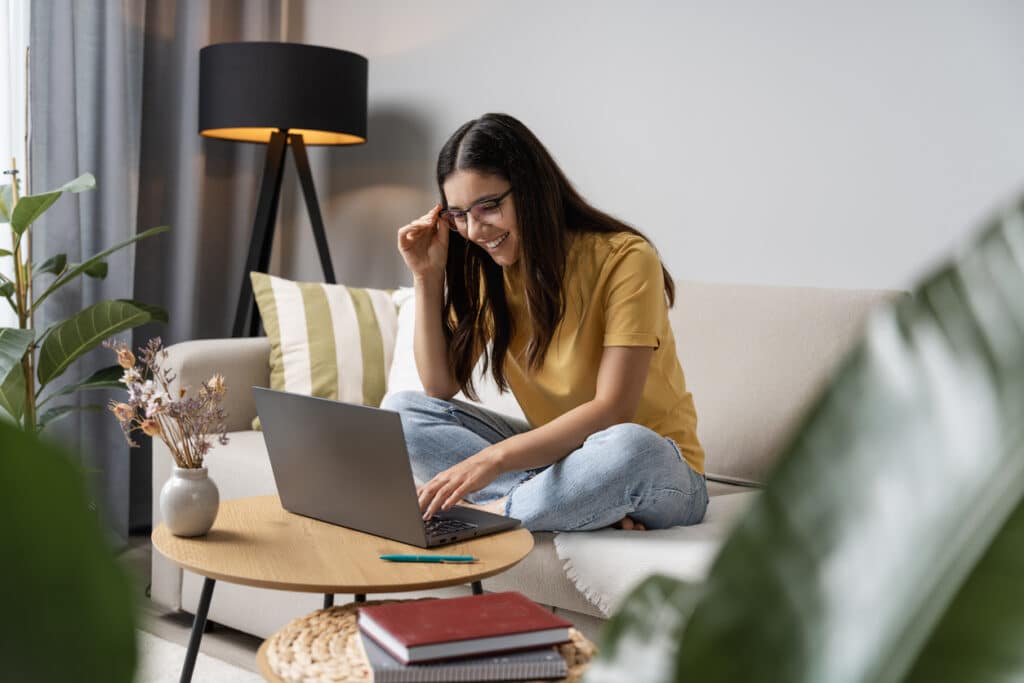 A person sitting cross‑legged on a beige sofa in a cozy living room, looking at a laptop on a round wooden coffee table. The scene includes a striped cushion, a black floor lamp, potted plants, dried flowers in a vase, and books on the lower shelf of the table, creating a warm and relaxed planning atmosphere.