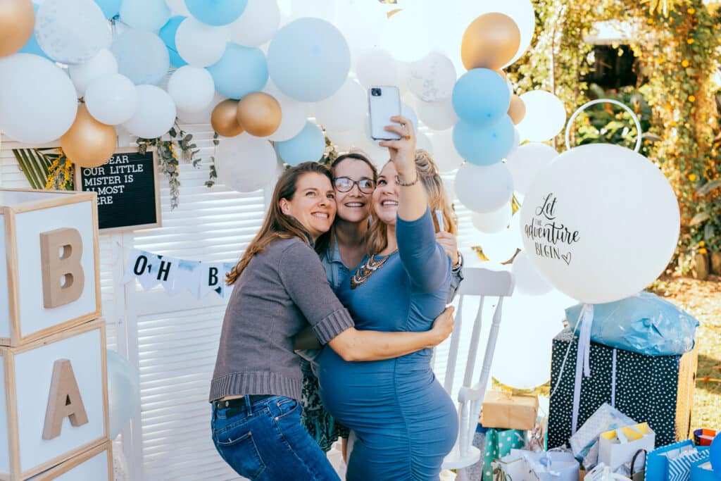Three guests taking a selfie at an outdoor baby shower, standing in front of a blue‑and‑gold balloon backdrop with “Oh Baby” signage, BABY blocks, wrapped gifts, and a large balloon reading “Let the adventure begin.