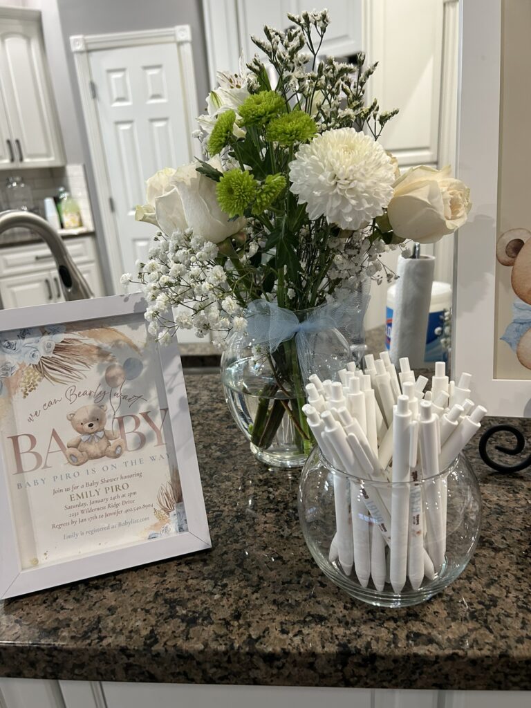 A framed baby shower invitation displayed on a counter with a floral arrangement and pens for a We Can Bearly Wait baby shower.