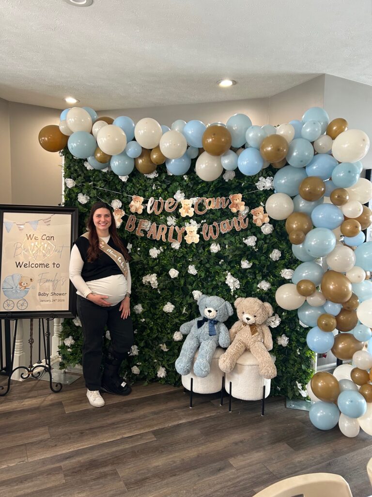 Mom‑to‑be standing in front of a We Can Bearly Wait baby shower backdrop with balloons, teddy bears, and a themed welcome sign.