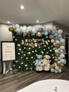 A “We Can Bearly Wait” baby shower photo booth setup with a greenery backdrop, pastel balloon garland, teddy bear banner, and two plush bears on stools.
