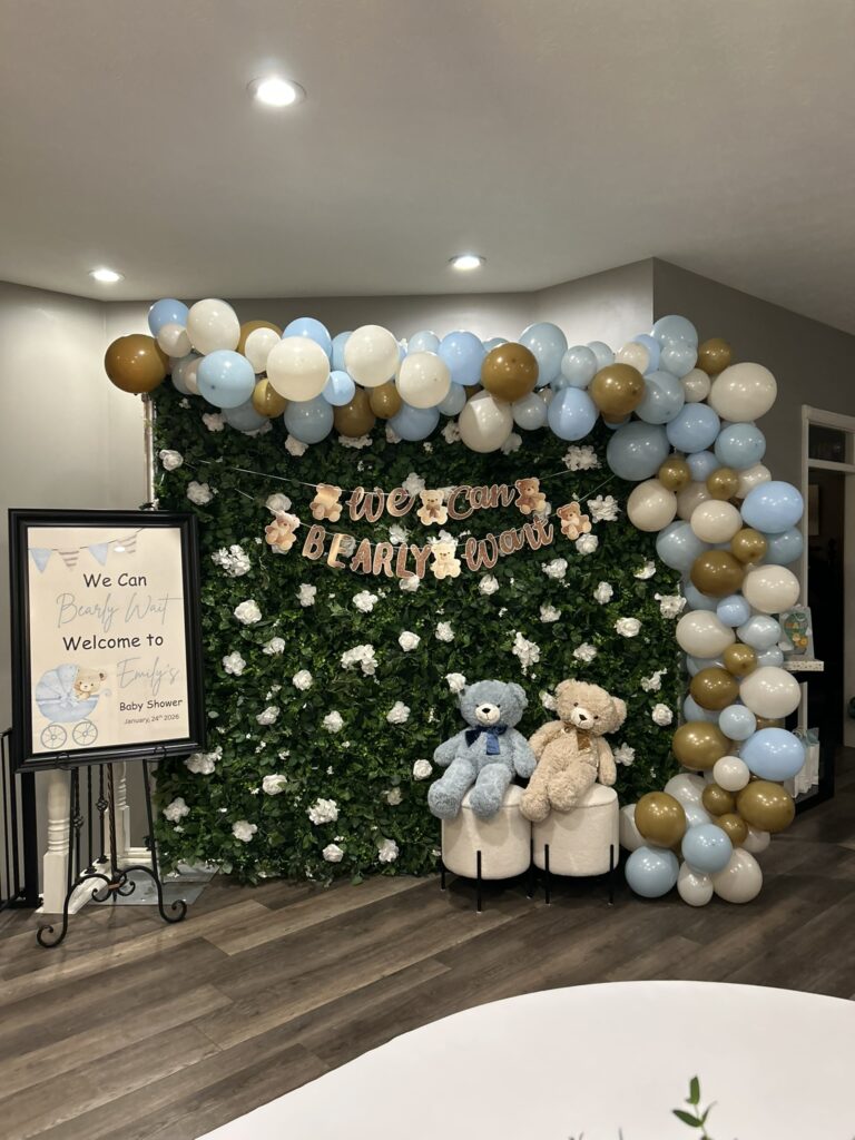 A “We Can Bearly Wait” baby shower photo booth setup with a greenery backdrop, pastel balloon garland, teddy bear banner, and two plush bears on stools.