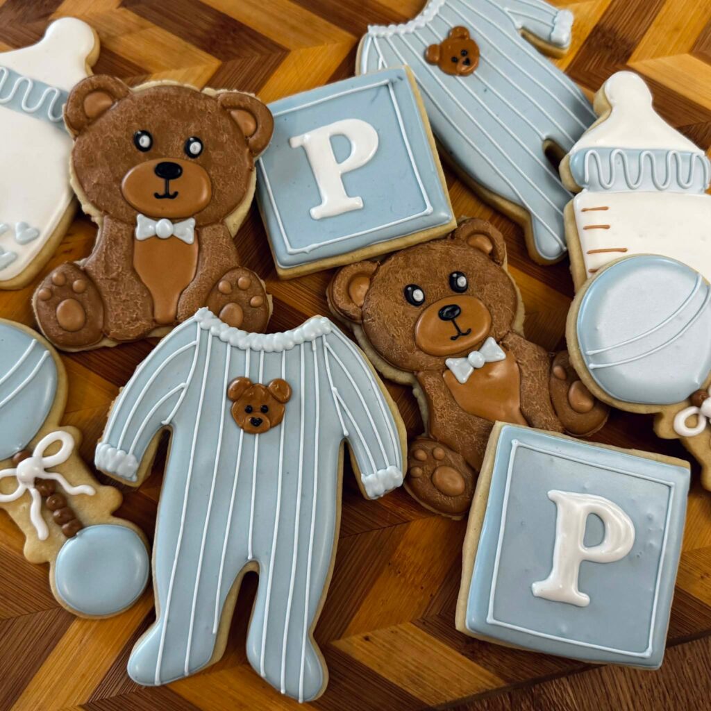 Decorated baby shower sugar cookies shaped like teddy bears, onesies, bottles, rattles, and letter blocks in soft blue, white, and brown for a We Can Bearly Wait baby shower.