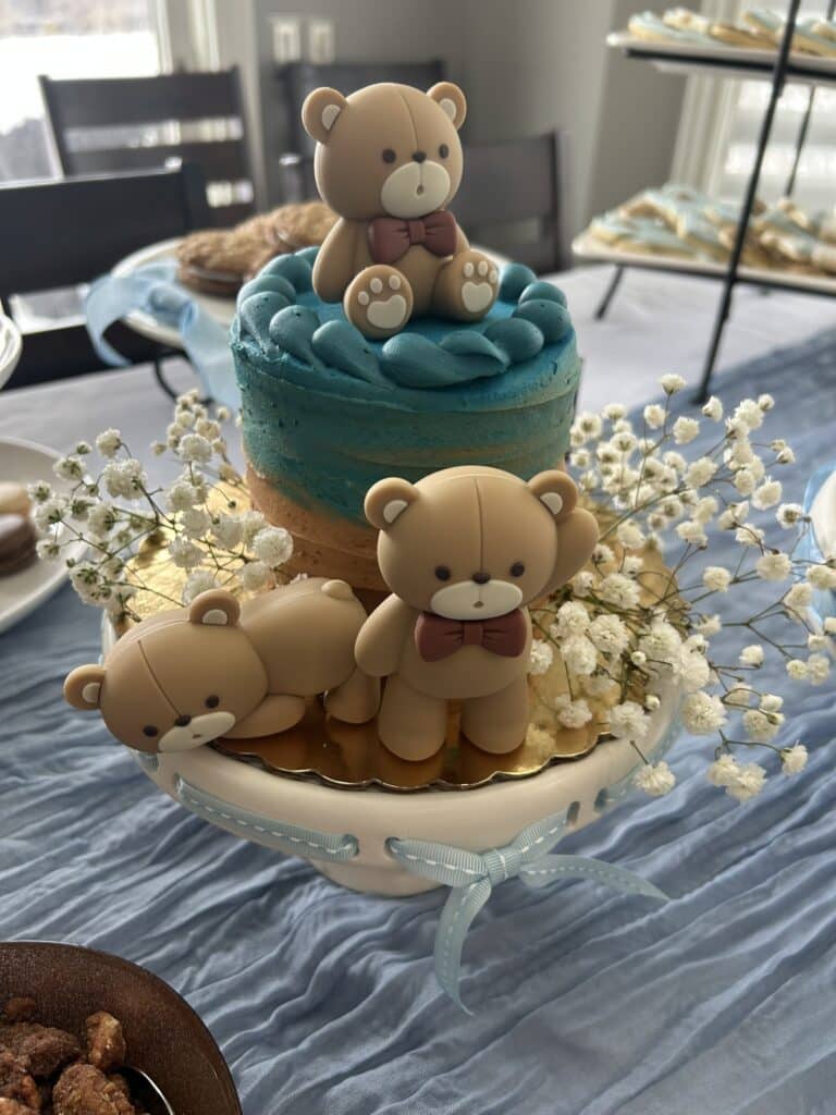 Blue and beige baby shower cake decorated with three fondant teddy bears and accented with small white flowers, with other themed desserts visible in the background.