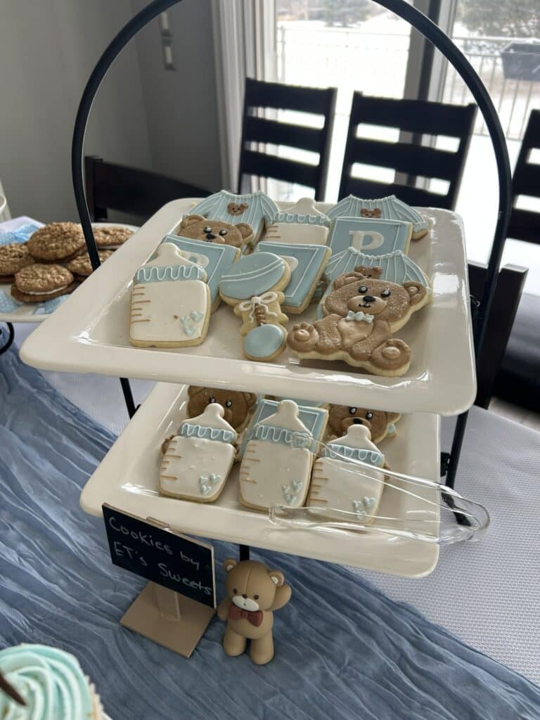 Tiered stand of baby‑themed decorated cookies, including onesies, bottles, bibs, pacifiers, and teddy bears, displayed with a small bear figurine and additional cookies in the background.