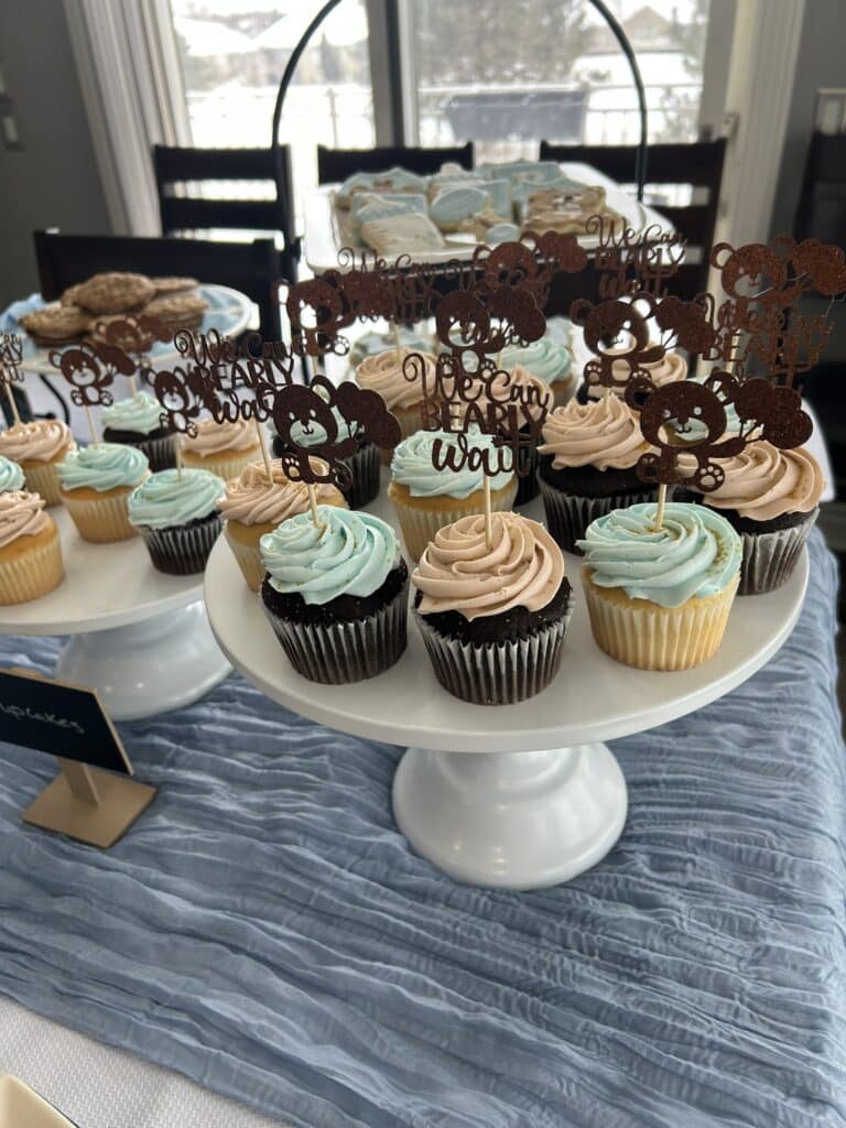 Cupcakes with pastel blue and beige frosting topped with “We can bearly wait” bear decorations, displayed on a white cake stand with additional themed cookies and treats in the background.