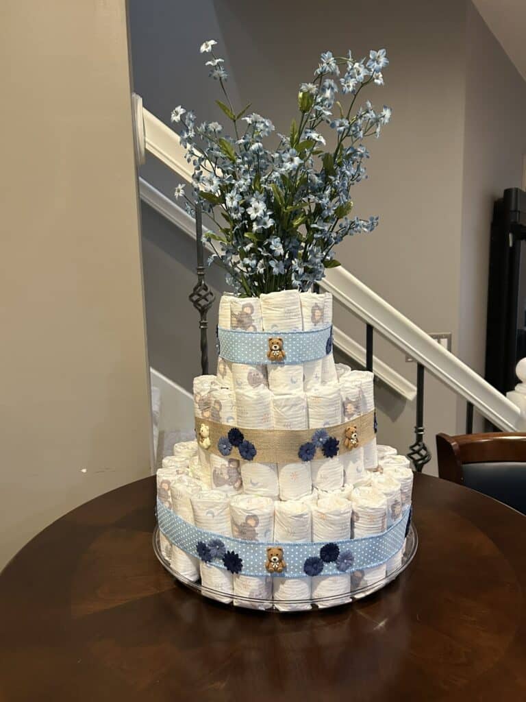 Three‑tiered diaper cake decorated with blue ribbons, burlap accents, teddy bear embellishments, and artificial blue flowers, displayed on a silver tray.