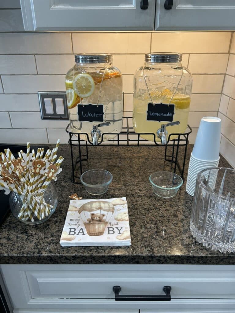 Beverage station with glass dispensers of citrus water and lemonade, stacked cups, bear‑themed paper straws, and napkins featuring a teddy bear in a hot air balloon.
